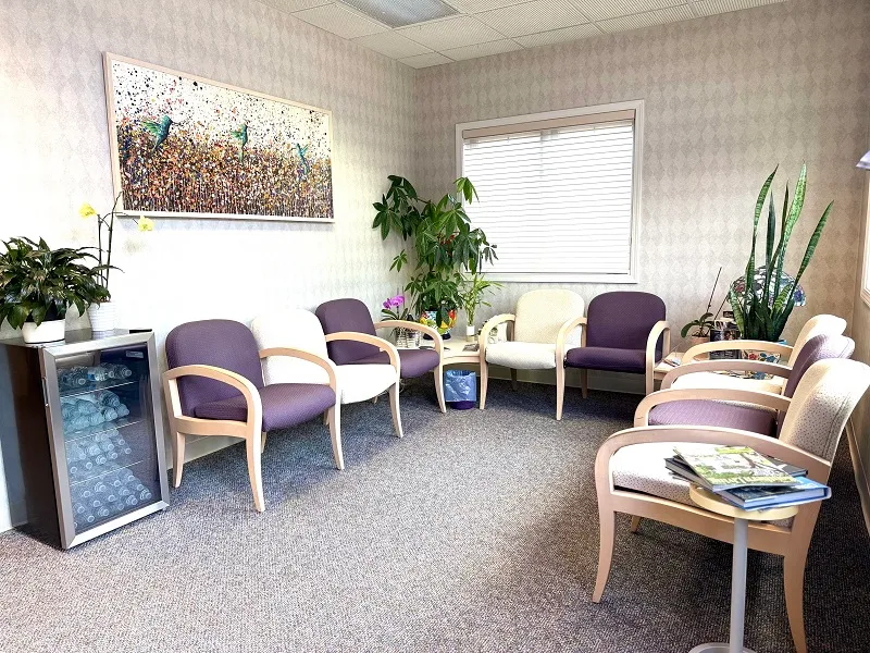 A cozy dental waiting room with comfortable purple and beige armchairs, indoor plants, and a small refrigerator for refreshments.