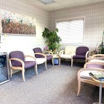 A cozy dental waiting room with comfortable purple and beige armchairs, indoor plants, and a small refrigerator for refreshments.