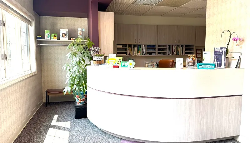 A bright dental reception area with a curved white front desk, large windows, and a purple accent wall in the background.