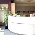 A bright dental reception area with a curved white front desk, large windows, and a purple accent wall in the background.