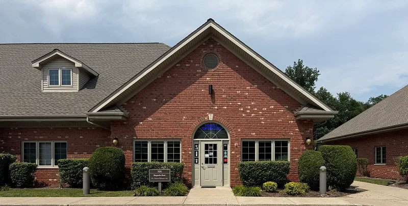 Exterior view of a professional red brick building with a gabled roof, featuring a central arched doorway and manicured green bushes.