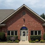 Exterior view of a professional red brick building with a gabled roof, featuring a central arched doorway and manicured green bushes.