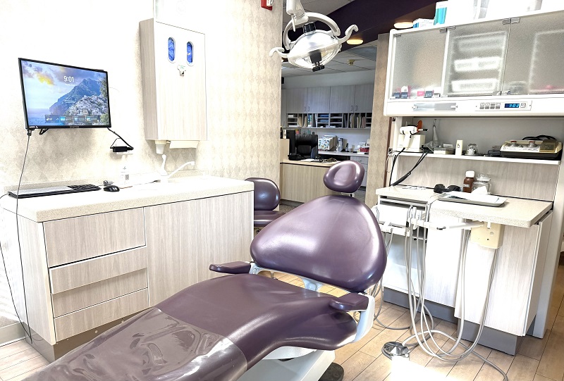 A close-up of a dental operatory featuring a purple reclining chair, overhead clinical lighting, and a wall-mounted monitor for patient education.